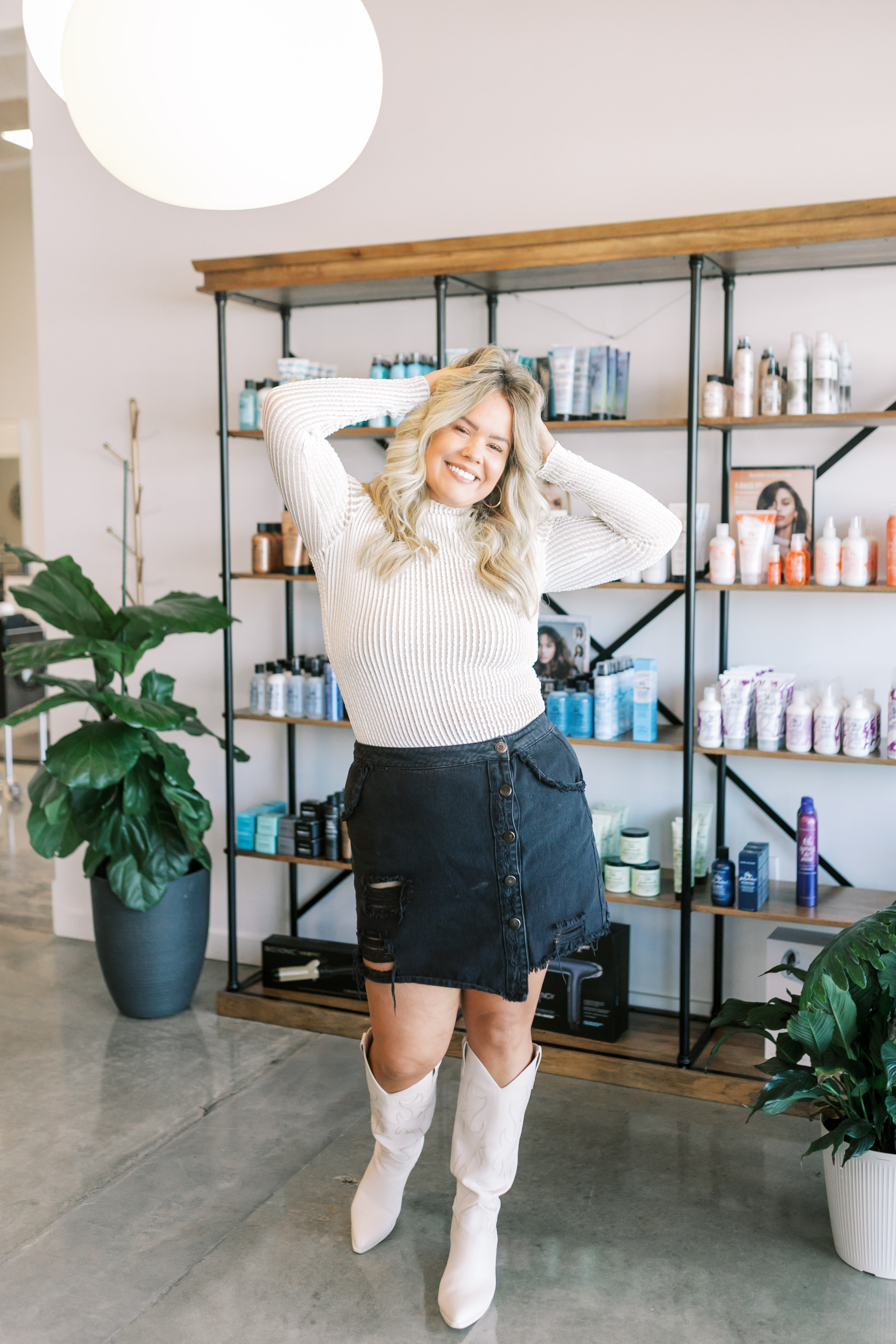 Stylist with blonde hair black skirt and white sweater posing with both hands in her hair